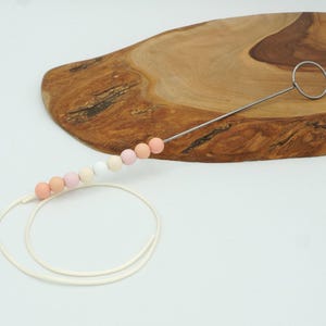 Puede incluir: Una cuerda blanca con una serie de cuentas de color rosa pastel, melocot&oacute;n y blanco enhebradas en un alambre de metal con un bucle en el extremo. Las cuentas est&aacute;n dispuestas en un degradado.