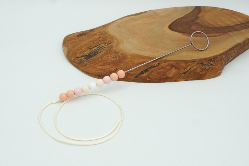 Puede incluir: Una cuerda blanca con una serie de cuentas de color rosa pastel, melocot&oacute;n y blanco enhebradas en un alambre de metal con un bucle en el extremo. Las cuentas est&aacute;n dispuestas en un degradado.
