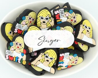 Puede incluir: Una colección de dijes de silicona negros, blancos, amarillos y multicolores que muestran una figura rubia con gafas de sol blancas. Cada dije muestra el texto 'little SWIFTIE' en letras coloridas, con pequeños detalles florales.