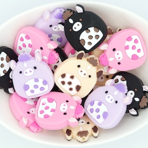 May include: A bowl filled with colorful silicone animal chew toys. The toys are shaped like cows and feature different colors, including pink, purple, black, and brown. Each toy has white spots and a cute face.