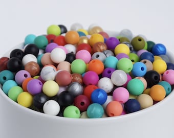 Puede incluir: Un bol lleno de cuentas de silicona de colores. Las cuentas son de varios tonos de azul, verde, rojo, naranja, amarillo, rosa, morado, marrón y gris. Las cuentas son todas redondas y tienen un agujero en el centro.