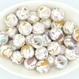 Peut inclure: Un bol de perles en céramique blanche avec un motif de champignons et de fleurs. Les perles mesurent environ 2 cm de diamètre et ont un fond blanc avec des accents bruns, jaunes, bleus et violets.