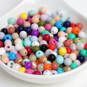 Peut inclure: Une collection de petites perles rondes multicolores dans un bol blanc en forme de goutte. Les perles présentent une variété de finitions unies, mates et métalliques dans des tons de rouge, bleu, vert, jaune, rose, violet, orange, noir, blanc et beige.