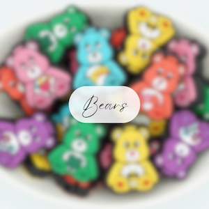 Peut inclure: Une collection de breloques d'ours de dessin animé colorés avec différentes expressions. Les breloques sont faites d'un matériau souple et malléable et présentent différentes couleurs, notamment le rose, le bleu, le jaune, le vert et le noir.