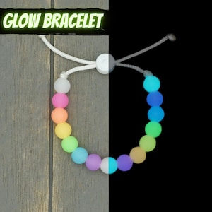 Puede incluir: Un brazalete blanco que brilla en la oscuridad con un arcoíris de colores. El brazalete está hecho de cuentas redondas y tiene una cuerda blanca.