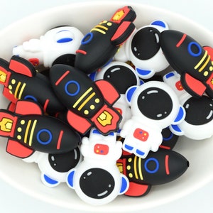 May include: A bowl filled with colourful silicone charms shaped like astronauts and rockets. The charms are black, white, red, blue and yellow.