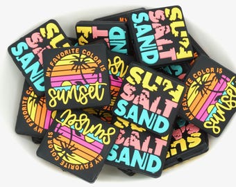 以下が含まれることがあります： カラフルな夕焼けのデザインと「SALT」、「SAND」、「Sunset」の文字が入った黒い正方形のチャームのコレクションです。チャームには「MY FAVORITE COLOR IS」の文字も入っています。
