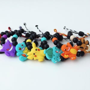 Puede incluir: Una colección de pulseras de cuentas de colores con dijes de personajes de dibujos animados. Las pulseras tienen cuentas negras y están adornadas con dijes en tonos morado, azul, amarillo, naranja y marrón. Los dijes son de personajes populares.