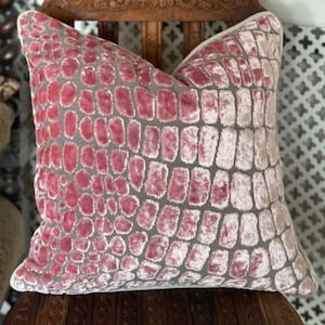 May include: A pink and grey patterned cushion with a subtle crocodile skin texture. The cushion has a white border and is sitting on a wooden chair.