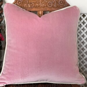 May include: A pink velvet cushion with a white border. The cushion is sitting on a wooden chair with intricate carvings.