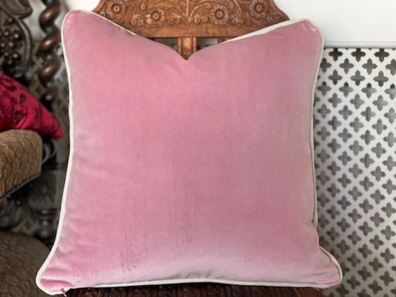 May include: A pink velvet cushion with a white border. The cushion is sitting on a wooden chair with intricate carvings.