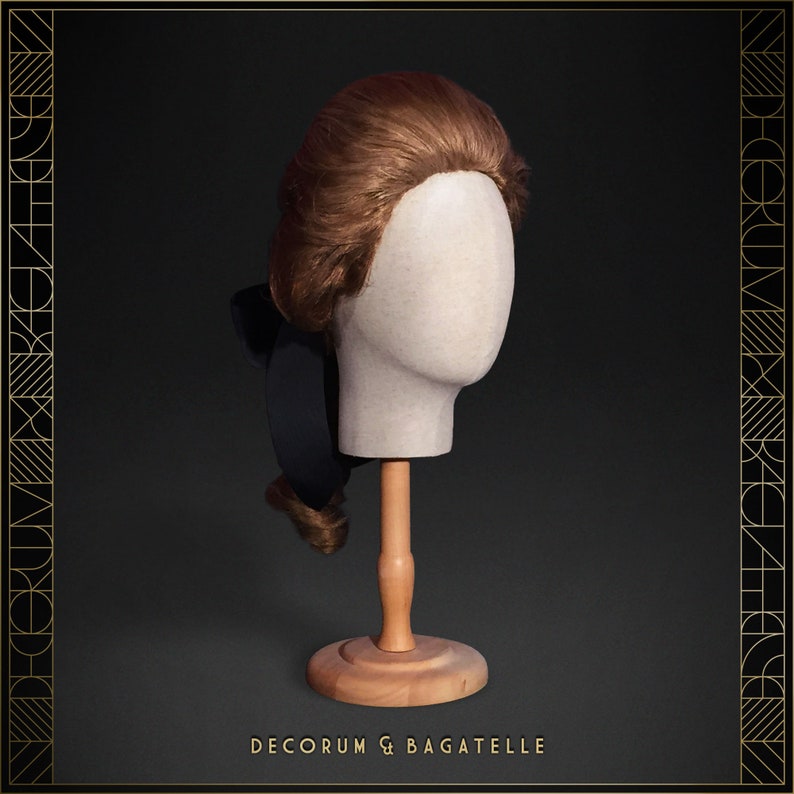 May include: A brown wig styled in a classic updo, displayed on a mannequin head. The wig features a dark bow and is mounted on a wooden stand. The background is dark with gold art deco style borders. The text "DECORUM & BAGATELLE" is at the bottom.
