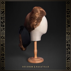 May include: A brown wig styled in a vintage updo, displayed on a mannequin head with a wooden stand. A black bow is attached to the side. The text "DECORUM & BAGATELLE" is at the bottom.