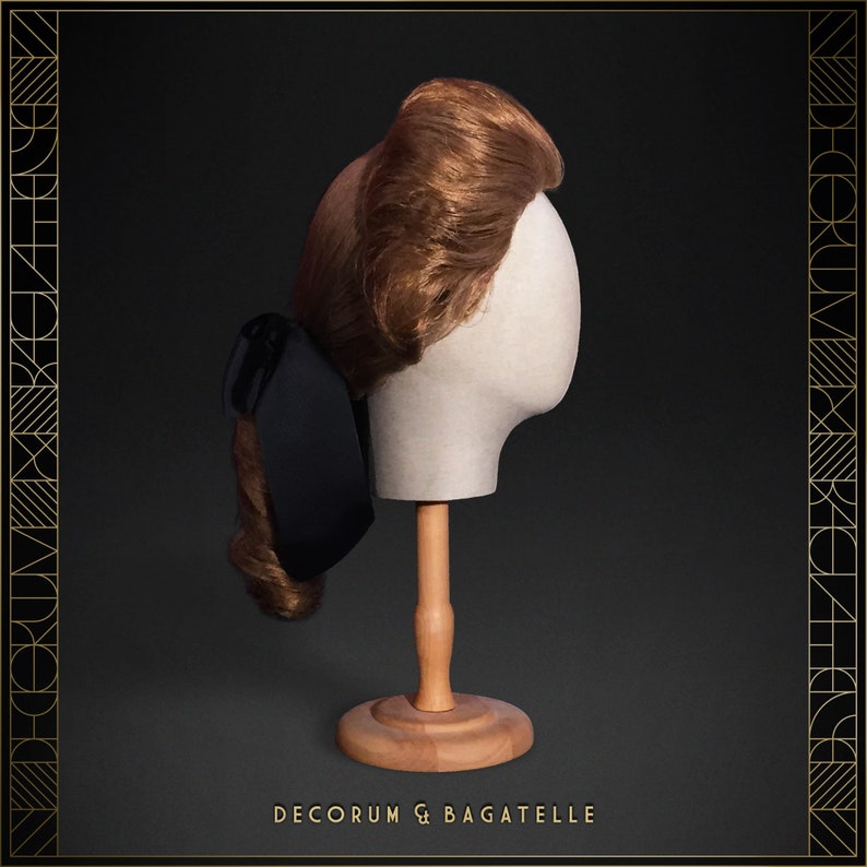 May include: A brown wig styled in a vintage updo, displayed on a mannequin head with a wooden stand. A black bow is attached to the side. The text "DECORUM & BAGATELLE" is at the bottom.