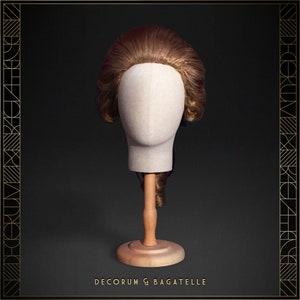 May include: A brown wig styled with curls and waves, displayed on a mannequin head. The head is mounted on a wooden stand. The background is dark with gold art deco style borders and the text "DECORUM & BAGATELLE".