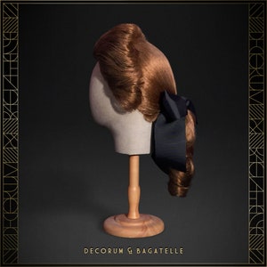 May include: A vintage-style auburn wig with curled ends and a dark blue bow, displayed on a mannequin head. The wig is styled with a side part and a voluminous top. The base is a light beige color, and the stand is made of wood. The text "DECORUM & BAGATELLE" is at the bottom.