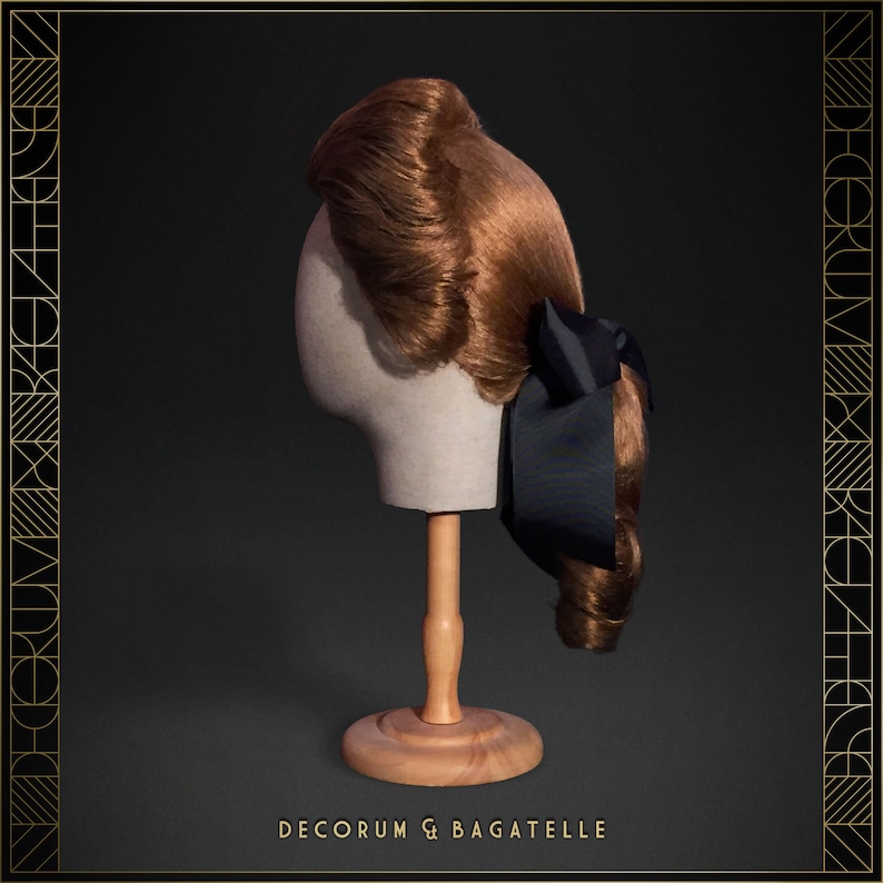 May include: A vintage-style auburn wig with curled ends and a dark blue bow, displayed on a mannequin head. The wig is styled with a side part and a voluminous top. The base is a light beige color, and the stand is made of wood. The text "DECORUM & BAGATELLE" is at the bottom.