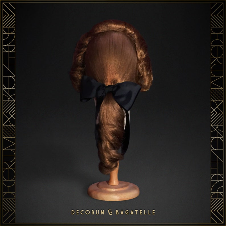 May include: A brown wig with a black bow, displayed on a wooden stand. The wig has a classic hairstyle with curls and a ponytail. The text "DECORUM & BAGATELLE" is at the bottom of the image.