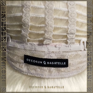 May include: Close-up of the interior of a wig, showing the construction with a lace front and adjustable straps. The wig features a light blonde color and a black label that reads "DECORUM & BAGATELLE". The bottom of the image also displays the text "DECORUM & BAGATELLE".
