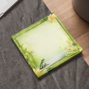 May include: A green frog sits on a green and yellow floral notepad with a white background. The notepad has a green border.