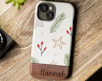 Könnte beinhalten: Eine weiße Handyhülle mit einem Weihnachtsmotiv mit grünen Tannenzweigen, roten Beeren und goldenen Sternen. Der untere Teil der Hülle ist in Holzoptik mit dem eingravierten Namen "Hannah" versehen.