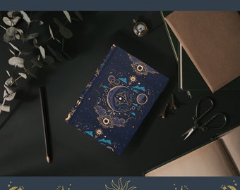 Op de afbeelding: Een donkerblauw notitieboek met een gouden en wit hemels ontwerp. Het ontwerp bevat een wassende maan, sterren en ogen. Het notitieboek ligt op een donker oppervlak met een potlood, een klein klokje en andere voorwerpen.