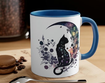 May include: A white ceramic mug with a blue rim and handle. The mug features a black silhouette of a cat sitting on a crescent moon with a galaxy design. The moon is surrounded by floral designs in shades of purple, pink, and blue.