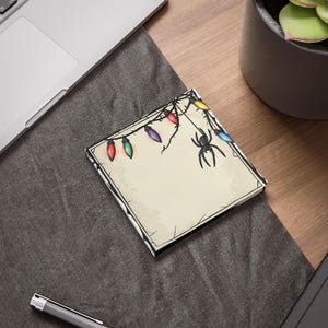May include: A square notepad with a spooky Halloween design. The notepad features a black spider hanging from a string of colorful Christmas lights.