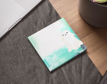 May include: A white ghost with a smiling face on a teal and white notepad with a watercolor background. The notepad has a white border and is on a gray surface.