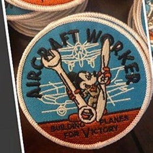 May include: A blue and white embroidered patch with a cartoon character holding two wrenches. The text on the patch reads "Aircraft Worker Building Planes For Victory".