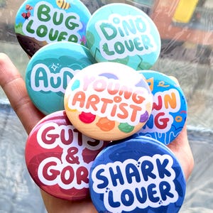 Peut inclure: Un ensemble de boutons ronds colorés avec différents motifs et textes. Les boutons présentent des phrases comme "Bug Lover", "Dino Lover", "Young Artist", "Guts & Gore" et "Shark Lover".