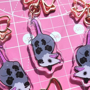 Op de afbeelding: Drie doorzichtige acryl sleutelhangers met een cartoon opossum met een grijs lichaam, zwarte pootjes en een roze staart. Elke sleutelhanger is bevestigd aan een ros&eacute;gouden metalen sluiting in de vorm van een strik.