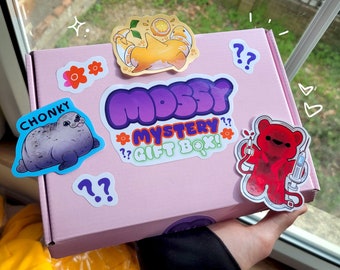 May include: A pink box with the text "Mossy Mystery Gift Box!" in purple, green, and white lettering. The box has various stickers on it, including a seal, a bear, and a cartoon character.