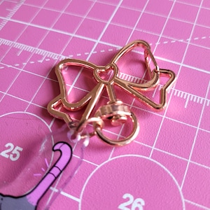 Op de afbeelding: Een ros&eacute;gouden metalen sleutelhanger in de vorm van een strik met een kleine hartvormige uitsparing in het midden. De sluiting heeft een draaimechanisme en een ring die is bevestigd aan een doorzichtige acryl hanger met een paars ontwerp.