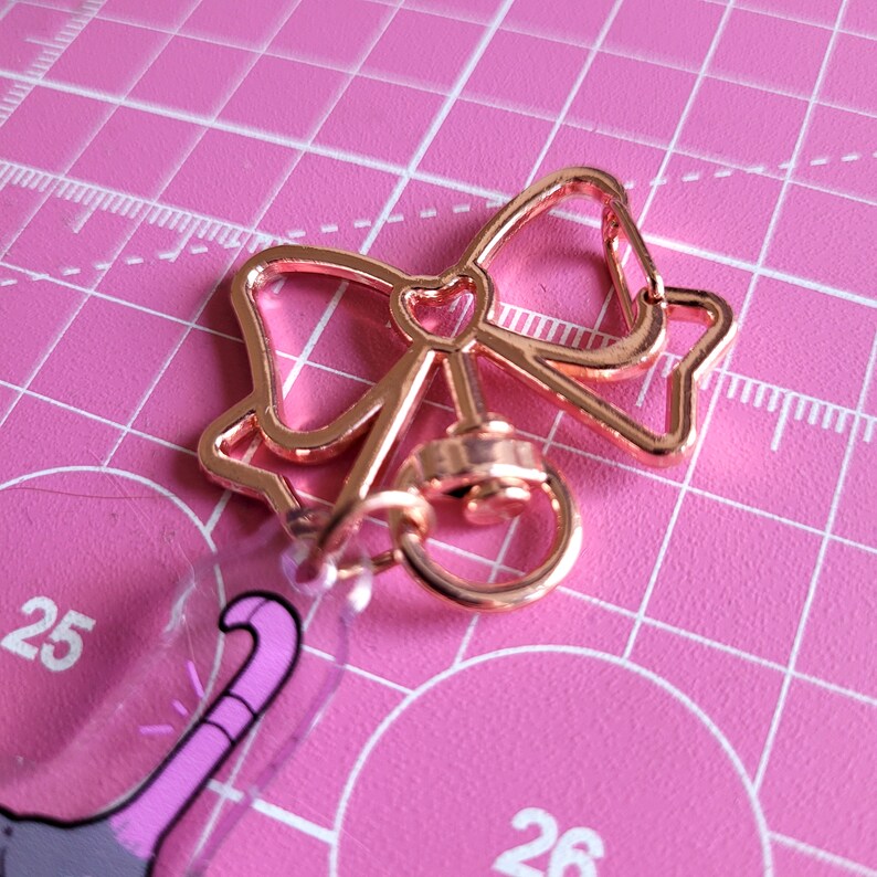Op de afbeelding: Een ros&eacute;gouden metalen sleutelhanger in de vorm van een strik met een kleine hartvormige uitsparing in het midden. De sluiting heeft een draaimechanisme en een ring die is bevestigd aan een doorzichtige acryl hanger met een paars ontwerp.