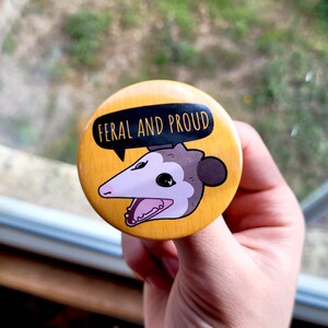 Puede incluir: Un bot&oacute;n amarillo con un opossum de dibujos animados con el texto "Feral and Proud" en negro.