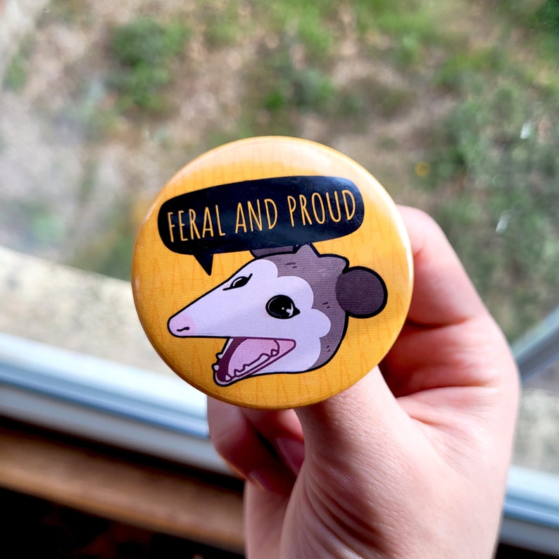 Puede incluir: Un bot&oacute;n amarillo con un opossum de dibujos animados con el texto "Feral and Proud" en negro.