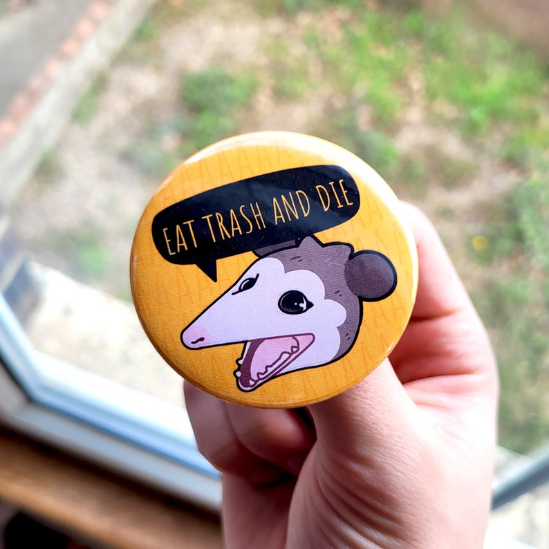 Puede incluir: Bot&oacute;n amarillo con un opossum de dibujos animados con el texto "Eat Trash and Die" en un bocadillo de di&aacute;logo.