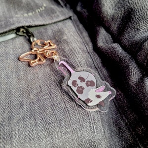 Op de afbeelding: Een doorzichtige acryl sleutelhanger met een cartoon opossum met een grijs lichaam, wit gezicht en roze accenten, bevestigd aan een ros&eacute;gouden vlindervormige metalen sluiting. De opossum is liggend afgebeeld met de bek open.