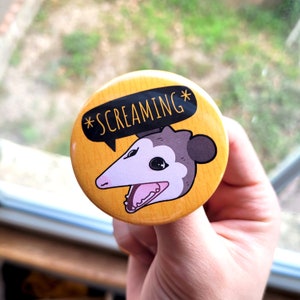 Puede incluir: Un bot&oacute;n amarillo con un opossum de dibujos animados con el texto "*SCREAMING*" en negro.