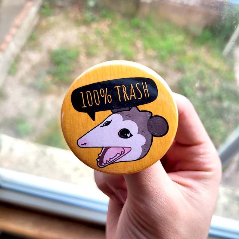 Puede incluir: Un bot&oacute;n amarillo con un opossum de dibujos animados con el texto "100% Trash" en &eacute;l.