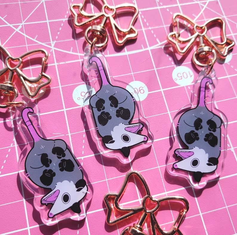 Op de afbeelding: Drie doorzichtige acryl sleutelhangers met een cartoon opossum met een grijs lichaam, zwarte pootjes en een roze staart. Elke sleutelhanger is bevestigd aan een ros&eacute;gouden metalen sluiting in de vorm van een strik.