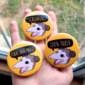 Puede incluir: Tres pines de bot&oacute;n amarillos con un opossum de dibujos animados en cada uno. Los opossums tienen diferentes expresiones y cada pin tiene un texto diferente. El primer pin dice "*SCREAMING*". El segundo pin dice "FERAL AND PROUD". El tercer pin dice "100% TRASH".
