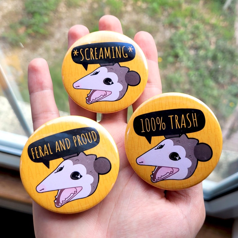 Puede incluir: Tres pines de bot&oacute;n amarillos con un opossum de dibujos animados en cada uno. Los opossums tienen diferentes expresiones y cada pin tiene un texto diferente. El primer pin dice "*SCREAMING*". El segundo pin dice "FERAL AND PROUD". El tercer pin dice "100% TRASH".