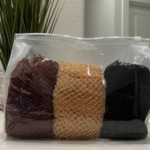 May include: A clear plastic bag containing three mesh bath sponges in shades of brown, tan, and black. The sponges are tightly packed and appear to be made of a woven material. The bag is sealed with a zipper.