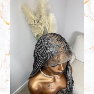 May include: A silver and black braided wig with a lace front is displayed on a mannequin head. The wig features intricate braiding patterns and a natural-looking hairline. The brand name "DW DAINTY WIGS" is visible on the mannequin.