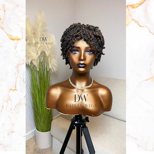 May include: A brown and blonde short curly wig on a mannequin head. The wig is styled in a twist out and is on a tripod stand. The text "DSW DAINTY WIGS" is on the stand.