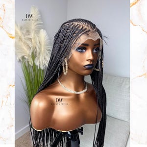 May include: A black braided wig displayed on a mannequin head. The wig has a lace front and is styled with small, neat braids. The mannequin is wearing gold hoop earrings and a diamond necklace. The brand name "DAINTY WIGS" is visible on the mannequin.