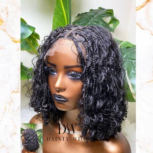Puede incluir: Una peluca rizada negra con detalles trenzados y una parte delantera de encaje. La peluca presenta un corte corto estilo bob con una línea de cabello de aspecto natural. La imagen incluye una imagen más pequeña de la peluca y el texto "DAINTY WIGS".