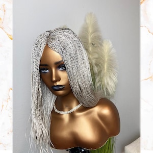 Op de afbeelding: Een zilver-zwarte gevlochten pruik op een mannequinhoofd. De pruik heeft een middenscheiding en lange, dunne vlechten. De mannequin heeft een goudkleurige buste en draagt een zilveren ketting. De achtergrond bevat decoratieve planten.